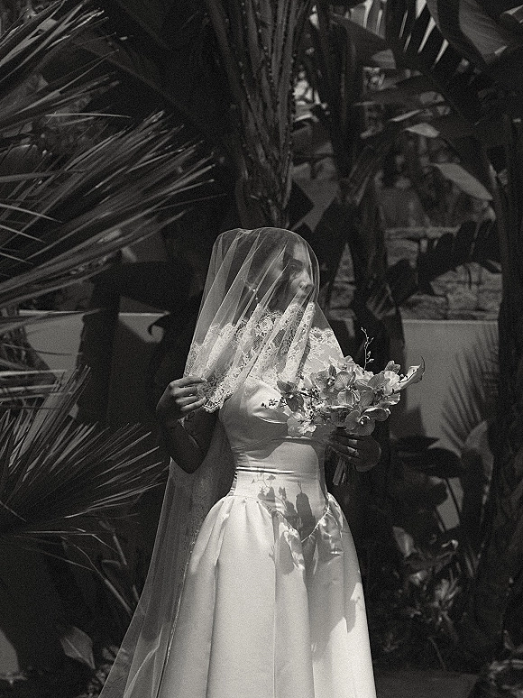 Bridal portrait of a bride holding bouquet with a lace-trim veil over her face, satin strapless gown, and palm-leaf garden backdrop