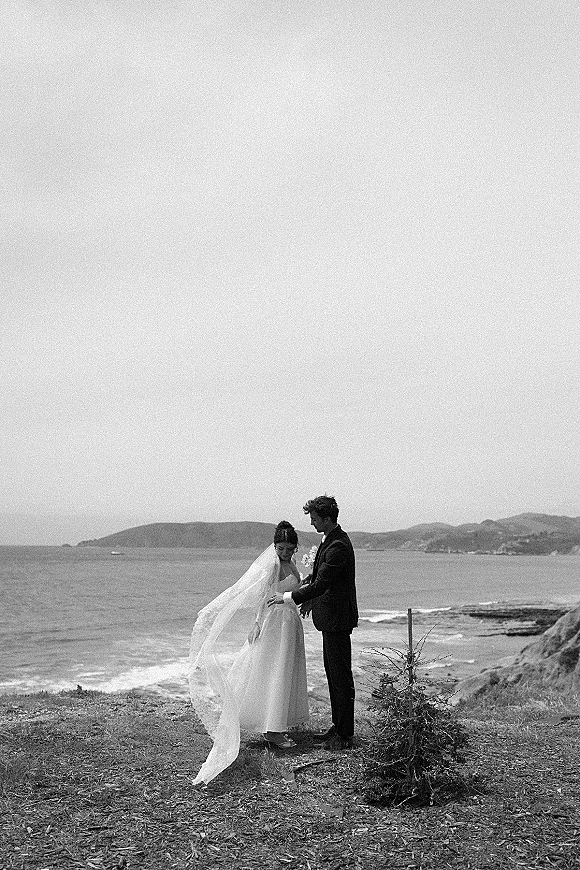 Couple portrait in a black and white wedding portrait style, bride with long veil and bouquet beside groom on a windy rocky coast with ocean backdrop