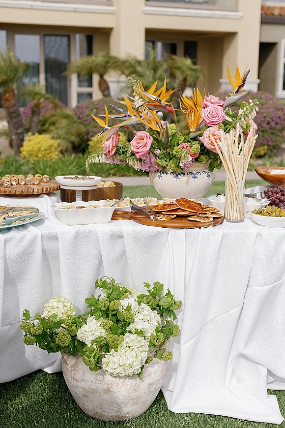 Wedding appetizer table with cocktail hour food table spread of breadsticks, crackers, grapes and dips, with tropical florals on a lawn by palm trees