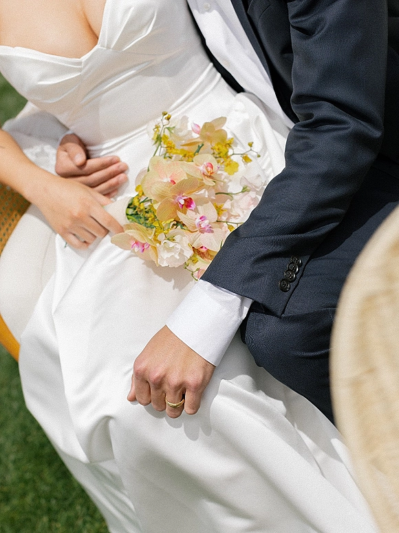 Couple portrait of bride and groom close up embracing, holding an orchid bridal bouquet against her satin gown on a green lawn in soft light