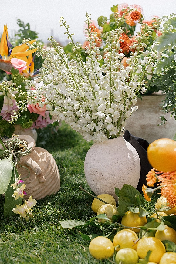 Wedding floral arrangement in a ceramic vase with white and orange blooms, greenery, and citrus fruit on a grass lawn outdoors