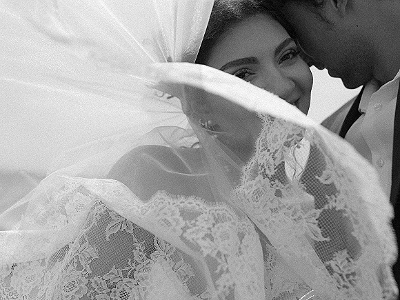 Wedding couple portrait of bride under veil as groom whispers cheek to cheek, lace detail and soft light against a bright sky