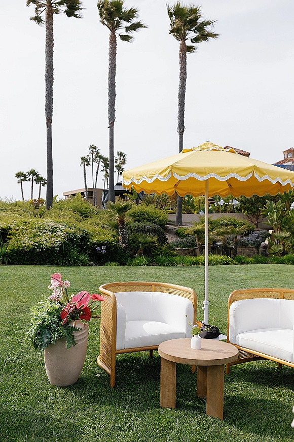 Outdoor lounge setup with wicker chairs and white cushions around a round wood table under a yellow umbrella on a palm-lined lawn