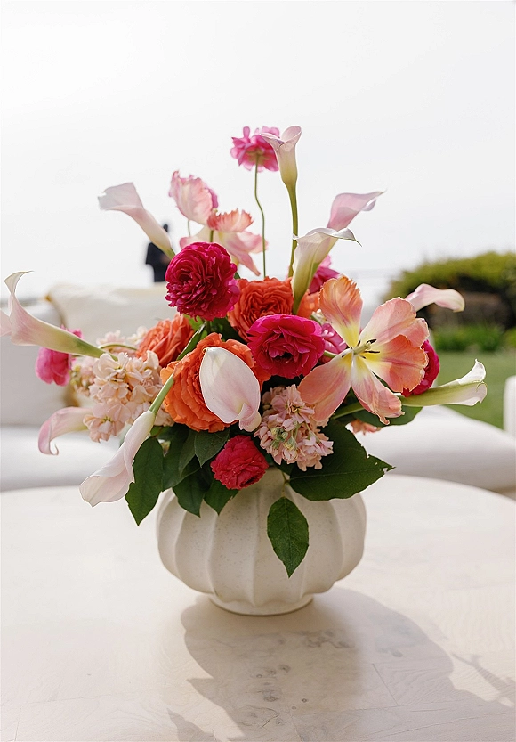 Wedding centerpiece of calla lilies, ranunculus and tulips in a white ceramic vase on a round table, with lawn and ocean view behind