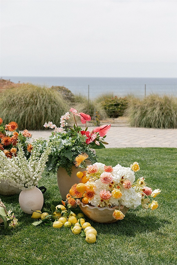 Wedding floral arrangement in a ceramic urn with hydrangeas, dahlias, anthurium, greenery, and citrus on a coastal lawn by dunes