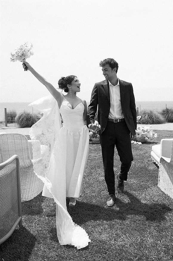 Newlywed couple walking hand in hand, bride holding bouquet as veil blows, on a grass lawn with open sky and horizon behind