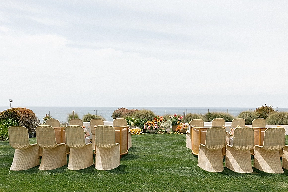 Ceremony setup with outdoor ceremony seating of wicker chairs lining a lawn aisle, grounded florals and citrus accents with ocean view beyond