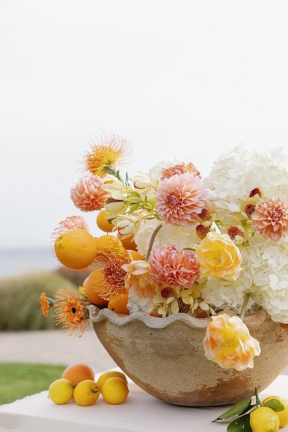 Wedding centerpiece with citrus wedding centerpiece blooms of hydrangea, dahlias, and roses in a stone bowl on an outdoor table against the sky