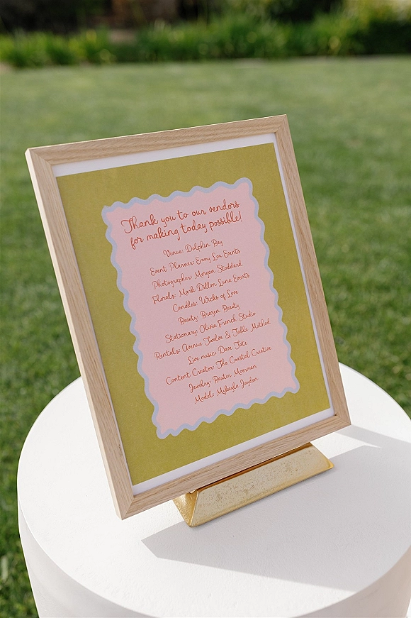 Wedding vendor sign in a wood frame with calligraphy lettering on a gold stand atop a round pedestal table on a green lawn