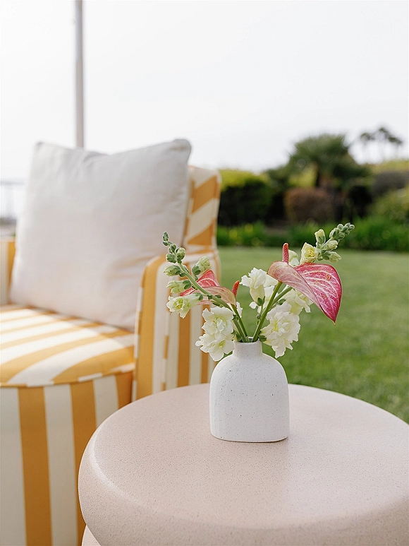 Wedding lounge seating with a striped armchair and white pillow beside a pedestal table with floral vase on a lawn overlooking the ocean