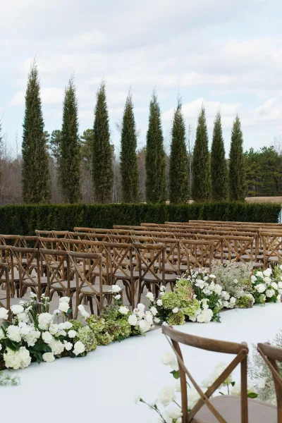 Ceremony aisle decor with a white aisle runner, crossback chairs, and white rose and hydrangea florals on a lawn beneath evergreens