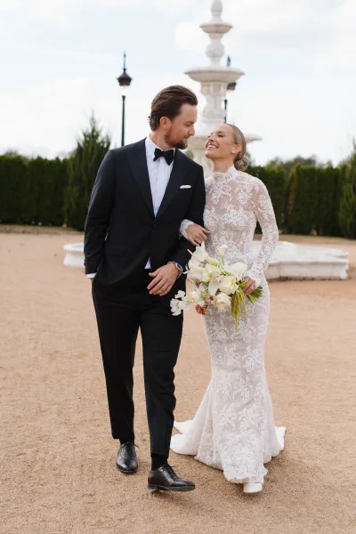 Couple portrait of bride and groom walking arm in arm, her bouquet in hand, by an outdoor fountain in a gravel courtyard with hedges