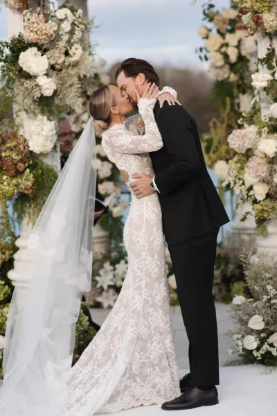 Wedding kiss as the bride in a lace dress and long veil kisses her tuxedoed groom beneath a white flower and greenery arch outdoors
