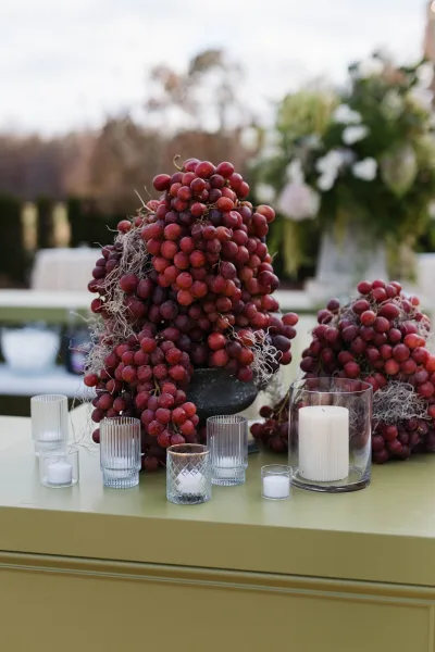 Reception centerpiece with red grapes around a pillar candle and white flowers in glass holders on an outdoor lawn table with trees behind