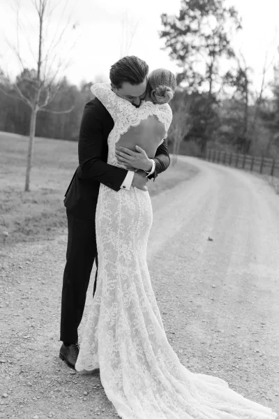 Wedding couple portrait in black and white, bride and groom hug on a gravel road, her low-back lace dress train and his tux visible near trees