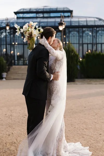Wedding kiss portrait of bride and groom kiss on stone steps, cathedral veil flowing, holding white flower bouquet by glass conservatory at dusk