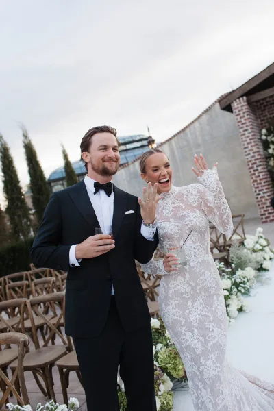 Ceremony moment as bride shows wedding ring while newlyweds laugh, holding cocktails in an outdoor brick courtyard aisle with white florals