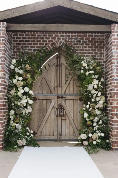 Ceremony altar with wedding floral arch of white flowers and greenery garland framing wooden doors against a brick wall entryway