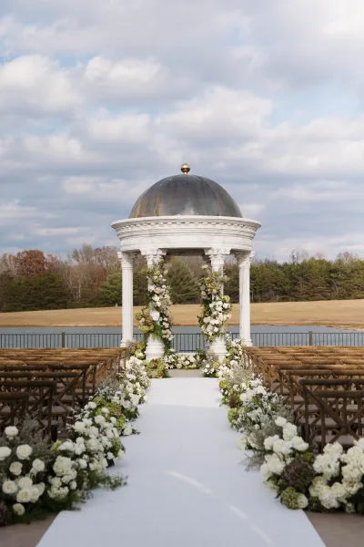 Ceremony aisle design with a white aisle runner flanked by ground florals, leading to a floral arch under a gazebo by the lake