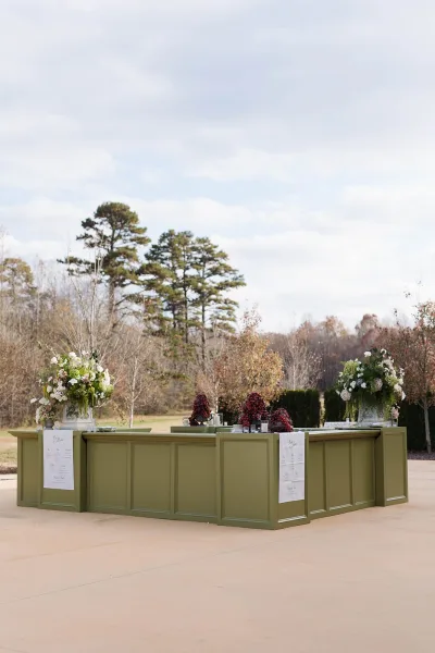 Wedding escort display with white calligraphy escort cards on olive green paneled stands, topped with flowers and grapes on an outdoor patio lawn