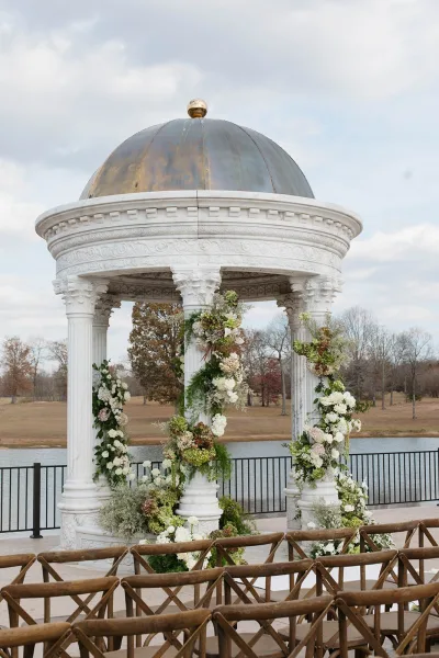 Wedding ceremony altar with floral arrangements and greenery garlands on a white gazebo, crossback chairs facing a lakeside view under cloudy sky