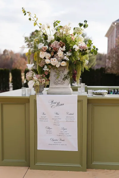 Wedding bar decor with a wedding bar menu sign on a green front panel, topped with white and blush flowers and glass votives on a lawn by a brick building