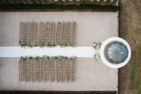 Ceremony setup with an outdoor ceremony layout featuring a white aisle runner, chair rows, and floral arrangements by a round fountain backdrop
