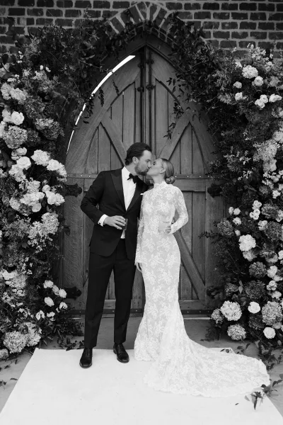 Wedding kiss portrait of bride and groom kissing under a rose and greenery floral arch at wooden church doors, bride holding a champagne coupe