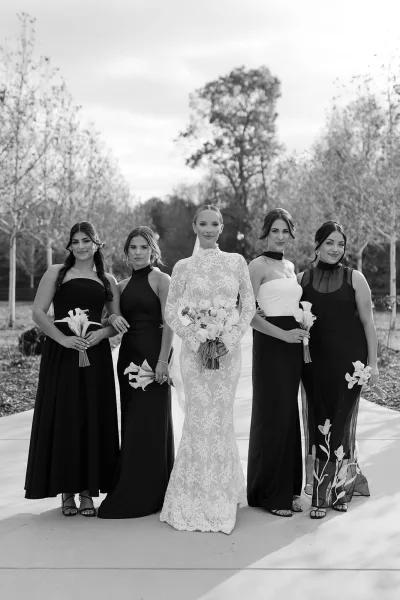 Bridal party portrait of bride with bridesmaids in mismatched black dresses, bride in long sleeve lace gown and veil on garden walkway