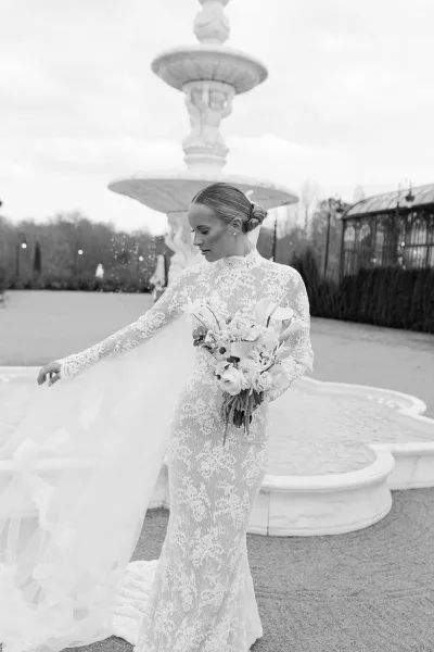 Bridal portrait in black and white of a bride holding bouquet of calla lilies and roses, veil blowing by a stone fountain in a courtyard