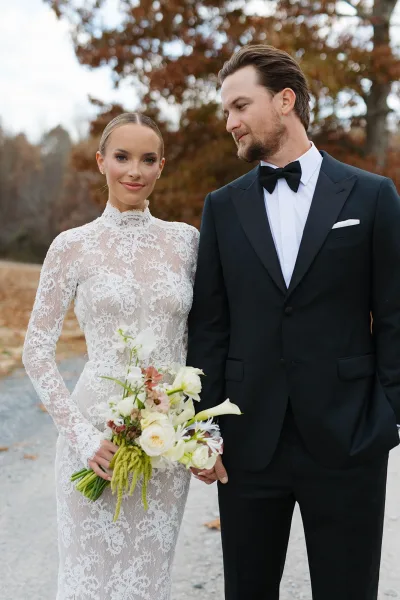 Couple portrait of bride in long sleeve high-neck lace gown holding white rose bouquet beside groom in black tux on autumn path