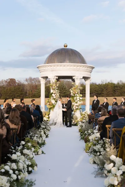 Ceremony moment as bride and groom exchange vows under a gazebo altar with floral arch, lakefront backdrop and seated guests