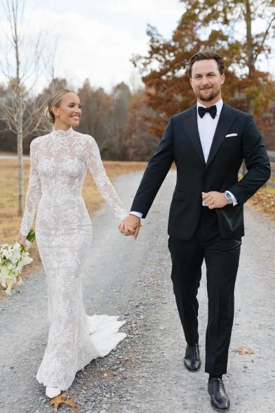 Couple portrait of bride and groom holding hands, walking a gravel road with autumn trees and fallen leaves, lace sleeves and bouquet visible