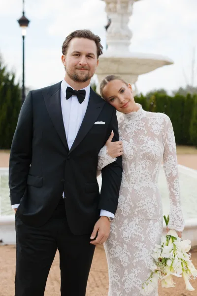 Couple portrait of bride and groom portrait by a stone fountain, bride in long-sleeve lace gown holding calla lily bouquet, groom in black tuxedo
