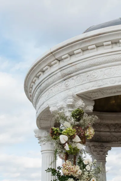 Wedding ceremony arch with hydrangeas and anthurium hanging florals, framed by ornate white columns in a carved stone pavilion under cloudy sky