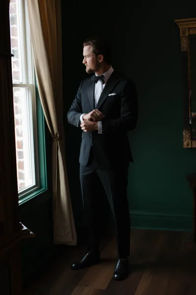 Groom portrait in tuxedo, a black tie groom adjusting cufflinks by a window, with dark green wall and mirror behind him