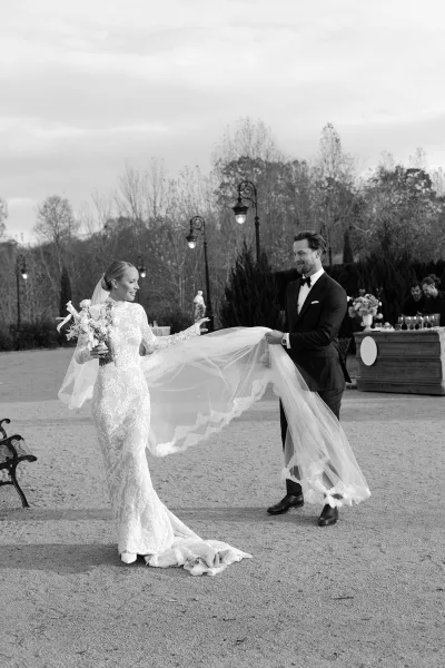 Couple portrait of bride and groom with veil on a garden gravel path, bride in lace dress holding white bouquet, groom in black tuxedo