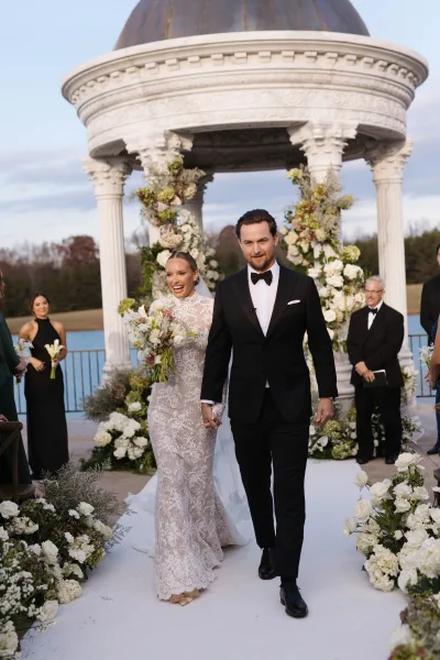 Recessional moment as bride and groom walk down aisle holding hands, passing white runner and florals under a gazebo by the lake