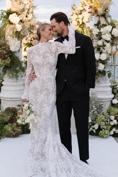 Couple portrait of bride and groom embrace with forehead touch, her lace long-sleeve gown and bouquet under a white rose arch in a garden with stone columns