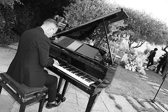 Wedding pianist plays a grand piano wedding setup with sheet music beside aisle flowers on a garden lawn as guests sit nearby