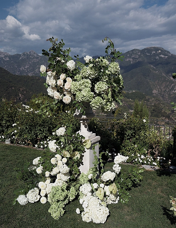 Wedding floral arrangement of white roses and hydrangeas in an urn on a vine-wrapped pedestal, set on a lawn with mountain views