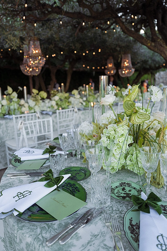 Reception tablescape with green and white tablescape florals, taper candles, crystal glassware and green napkin bows under garden string lights