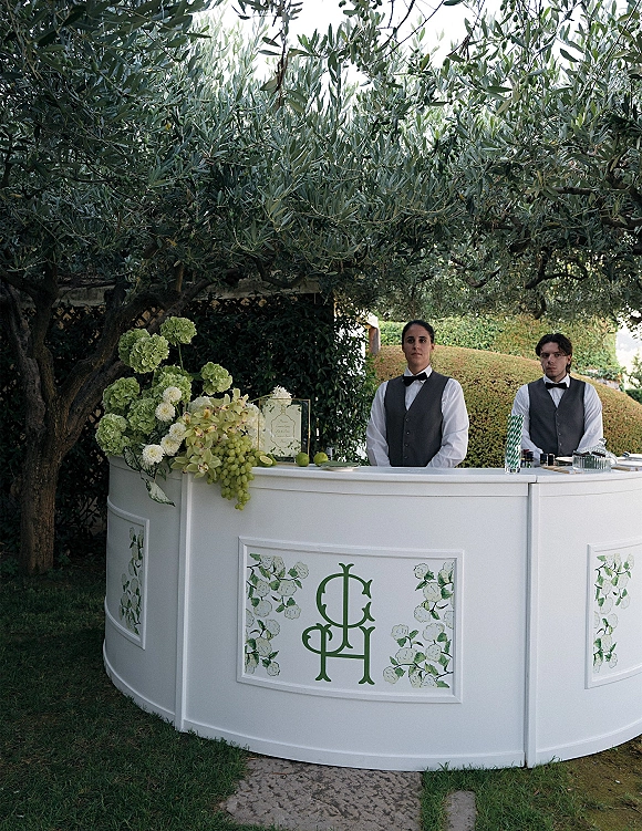 Wedding bar setup with a white curved bar, hydrangea flowers, grapes and limes, and bartenders in vests among olive trees and hedges