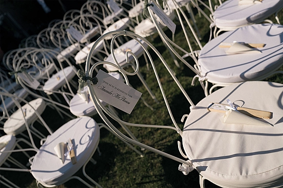 Ceremony seating with outdoor ceremony chairs, white wrought iron rows with reserved tags and ribbon-tied programs on a sunlit lawn