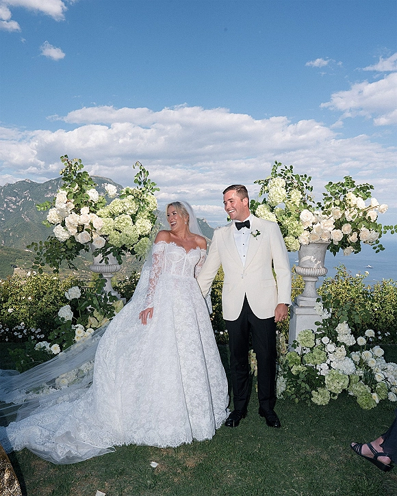 Couple portrait of bride and groom laughing, holding hands by a stone balustrade with mountains and lake behind, cathedral veil flowing