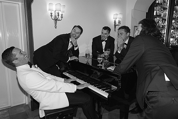 Groomsmen candid laughing in tuxedos as the groom in a white dinner jacket plays a grand piano at an indoor bar with champagne flutes