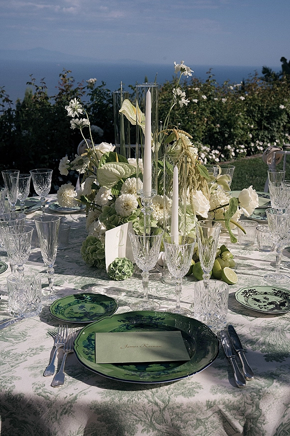 Reception tablescape with green and white tablescape place settings, crystal goblets, taper candles, and limes, ocean and mountains beyond hedges