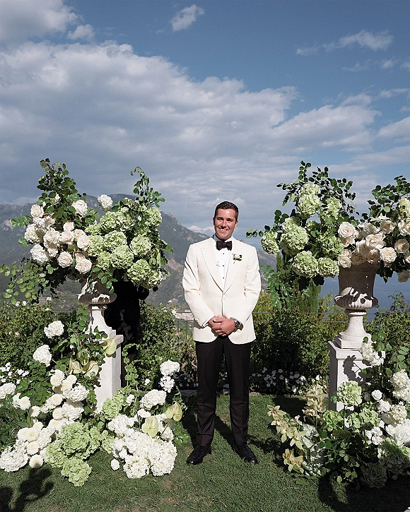 Groom portrait in a white tuxedo jacket with black bow tie and boutonniere, hands clasped in a garden with mountain view beyond