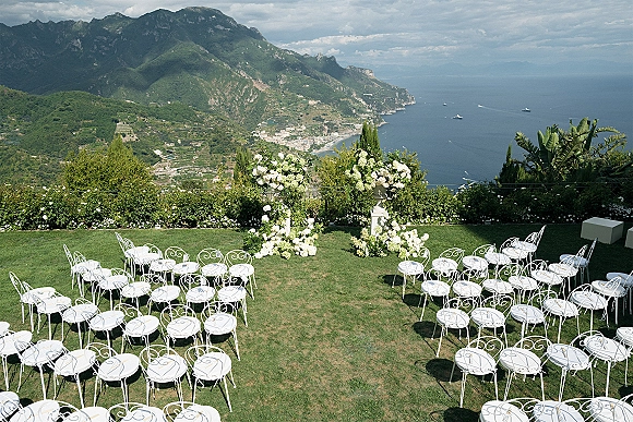 Ceremony setup with white floral arrangements on urn pedestals and wrought iron chairs arranged on a lawn overlooking the ocean and mountains