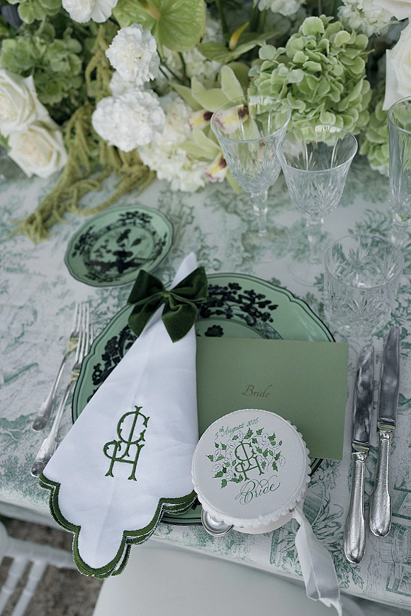 Reception place setting with green wedding tablescape featuring sage plates, monogrammed napkin with velvet bow, crystal glasses, white florals in outdoor light